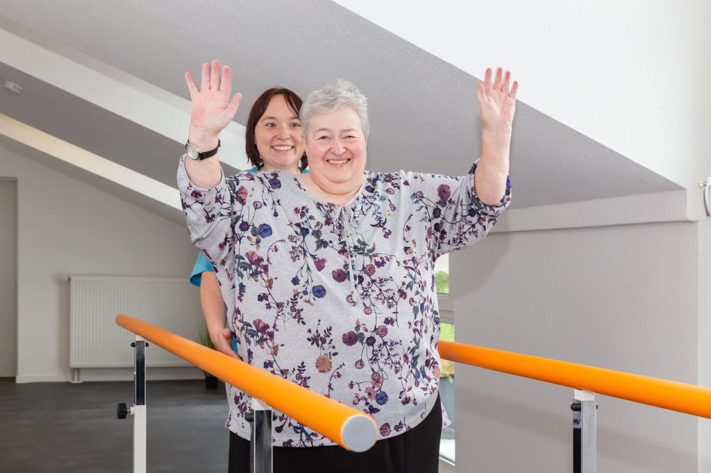 Eine Patientin der geriatrischen Reha steht mit erhobenen Händen freihändig an einem Trainingsbalken und freut sich über Ihren Fortschritt.